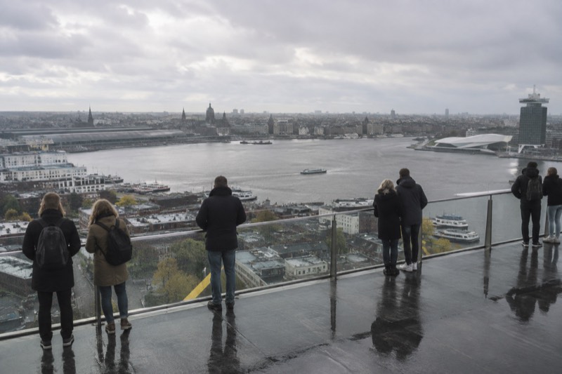 A rooftop observation deck high on a tower above a wide river, visitors at the glass railing looking out over low rooftops and harbour water, Amsterdam, Netherlands