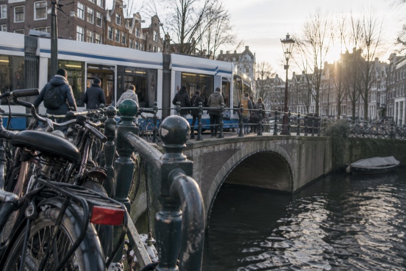 A city tram crossing a canal bridge with passengers stepping aboard at the stop and bicycles racked along the railing, Amsterdam, Netherlands