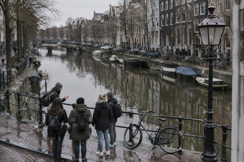 A guide talking to four visitors on a narrow pavement beside a canal, bicycles leaning on the railing and tall houses reflected in the water, Amsterdam, Netherlands
