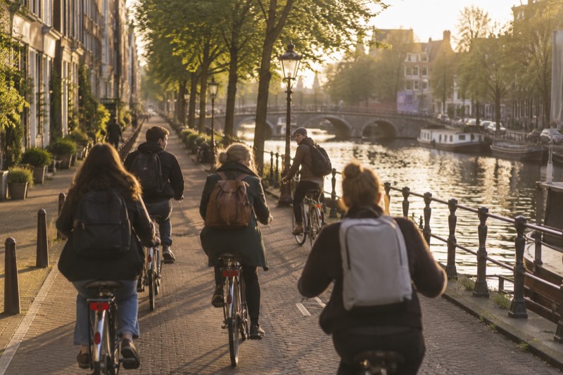 A group of cyclists on upright city bikes riding along a canal-side lane, the guide at the front turning to look back at them, Amsterdam, Netherlands