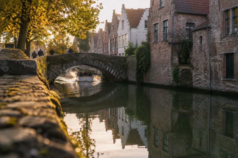 A quiet canal in the old centre of Bruges, Belgium, stepped brick gables reflected in still green water and a small open boat passing under a stone bridge
