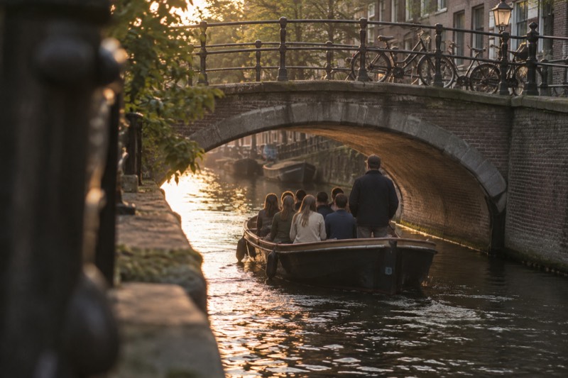 A small open canal boat carrying eight or nine passengers with a guide standing at the tiller, passing under a low arched brick bridge on a narrow canal in Amsterdam, Netherlands