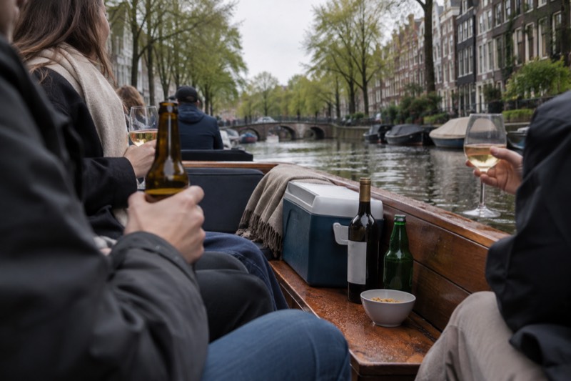 Passengers on an open-topped canal boat holding beer and wine glasses, bottles and a cooler on the bench beside them, a tree-lined canal sliding past, Amsterdam, Netherlands