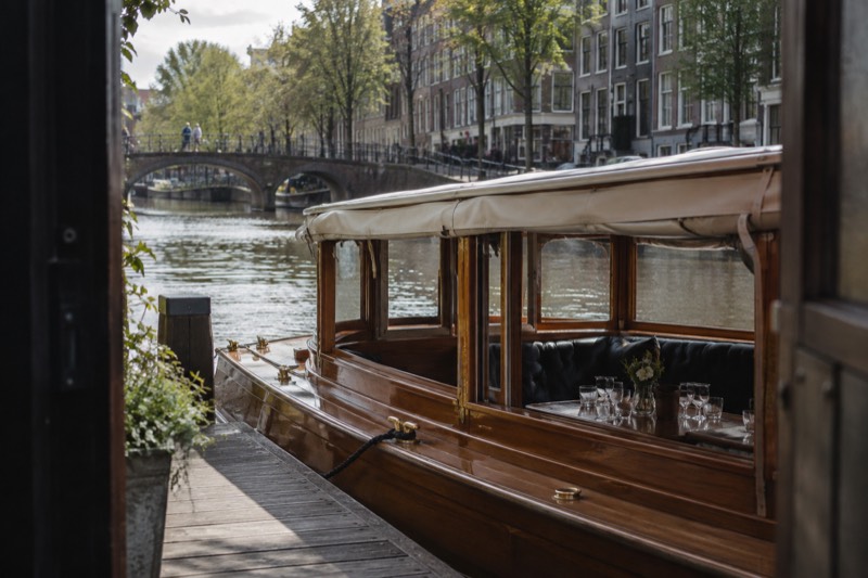 A varnished wooden saloon boat with brass fittings and a low canvas canopy moored on a quiet canal, glasses set out on the cabin table inside, Amsterdam, Netherlands
