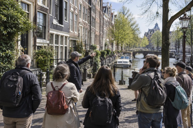 A guide gesturing toward a row of tall gabled merchant houses while a group of adults listens from the cobbles opposite, Amsterdam, Netherlands