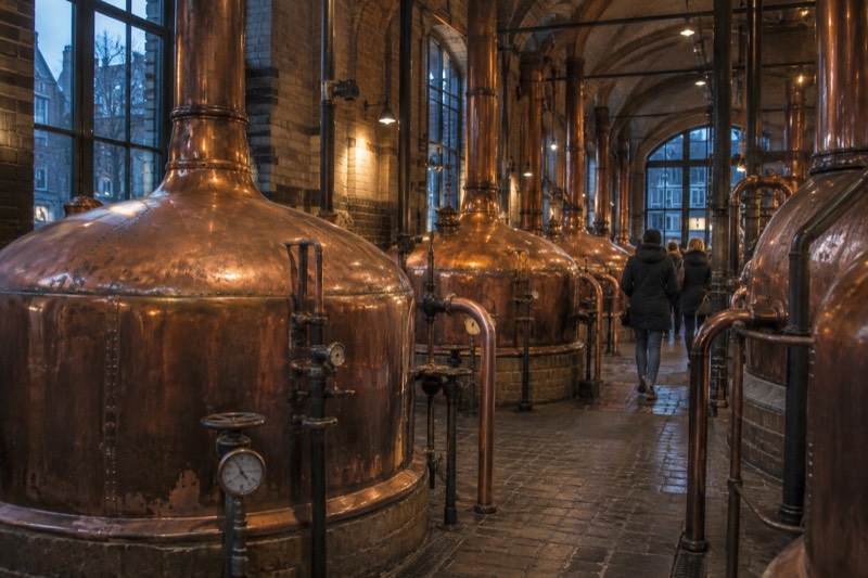 Rows of tall polished copper brewing kettles under a vaulted brick ceiling in a converted historic brewery, visitors walking between them, Amsterdam, Netherlands
