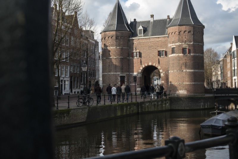 A group gathered beneath a squat medieval brick gatehouse with round corner towers at the edge of a canal, Amsterdam, Netherlands