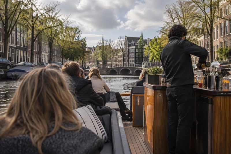 A modern low canal boat with a small bar at the stern, a crew member pouring a drink while passengers sit along the open sides, gabled houses behind, Amsterdam, Netherlands