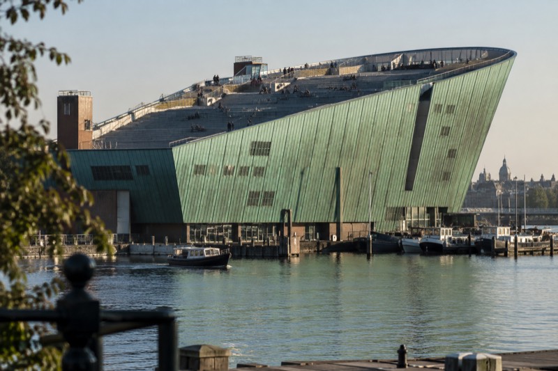 A huge green copper-clad building rising out of the water like a ship's prow, people sitting on its wide stepped roof terrace above the harbour, Amsterdam, Netherlands
