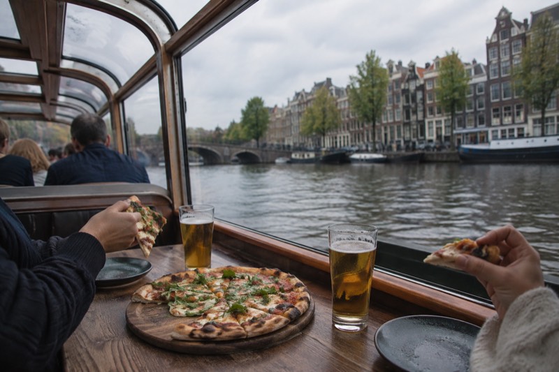 A table on a covered canal boat set with a whole pizza on a wooden board and glasses of beer, passengers eating as the canal slides past the window, Amsterdam, Netherlands