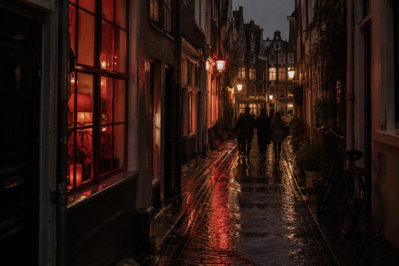 A narrow old lane in the medieval centre at night, warm and red window light reflected on wet cobbles, a small group walking away from the camera, Amsterdam, Netherlands