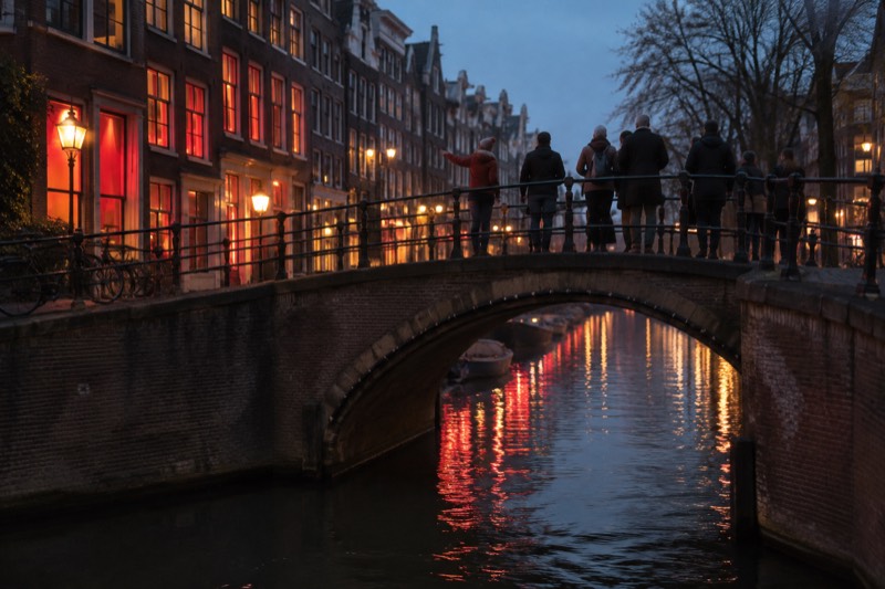 A guide and six visitors on an arched bridge over a narrow canal at night, red-tinted light lying on the water and the old brick facades, Amsterdam, Netherlands