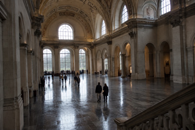 A vast vaulted museum hall with a high decorated ceiling and tall arched windows, visitors crossing the polished stone floor far below, bare walls, Amsterdam, Netherlands