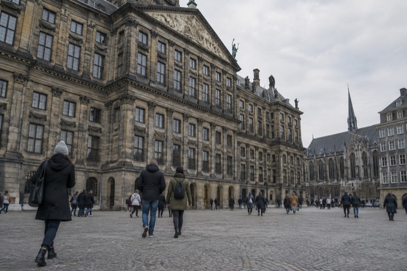 A monumental seventeenth-century sandstone palace with a heavy classical facade filling one side of a wide open square, people crossing the paving in front of it, Amsterdam, Netherlands
