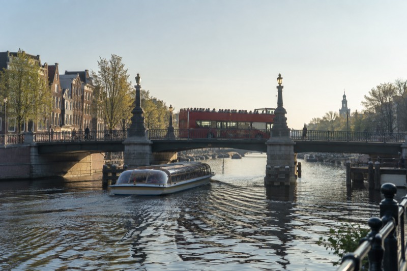 An open-topped double-decker sightseeing bus crossing a wide canal bridge with passengers on the upper deck, a canal boat passing beneath it, Amsterdam, Netherlands