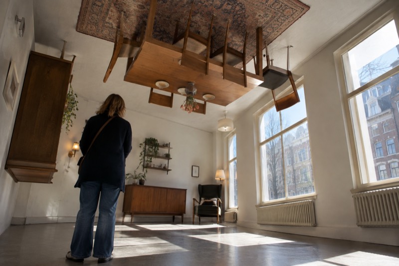 A brightly lit room built entirely upside down, a table and chairs bolted to the ceiling and a patterned rug above them, one visitor standing beneath, Amsterdam, Netherlands