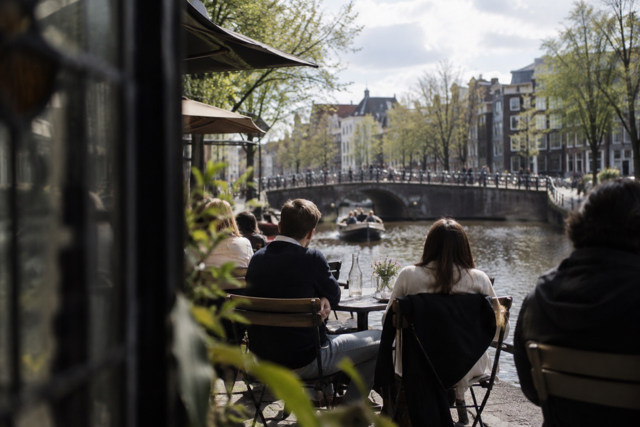 Canal-side restaurant terrace in Amsterdam with diners at small tables beside the water