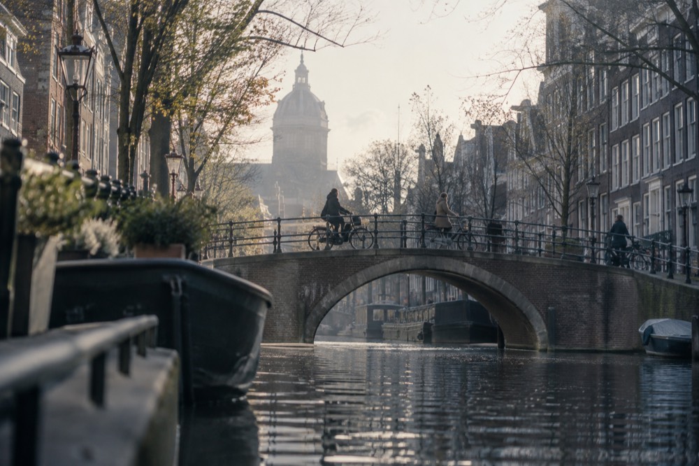 Bicycles crossing a small arched bridge over an Amsterdam canal lined with narrow brick houses