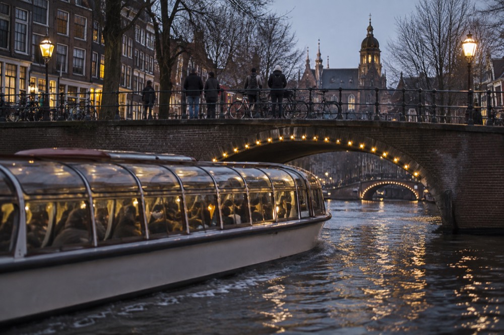 Enclosed canal cruise boat passing under a lit Amsterdam bridge on a winter evening