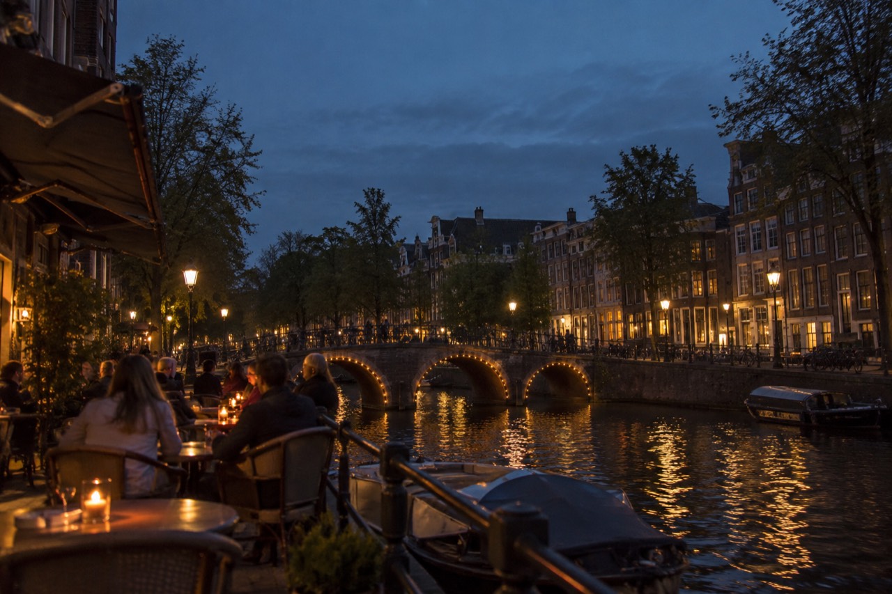 A canal side restaurant terrace with small tables and a bridge in the background