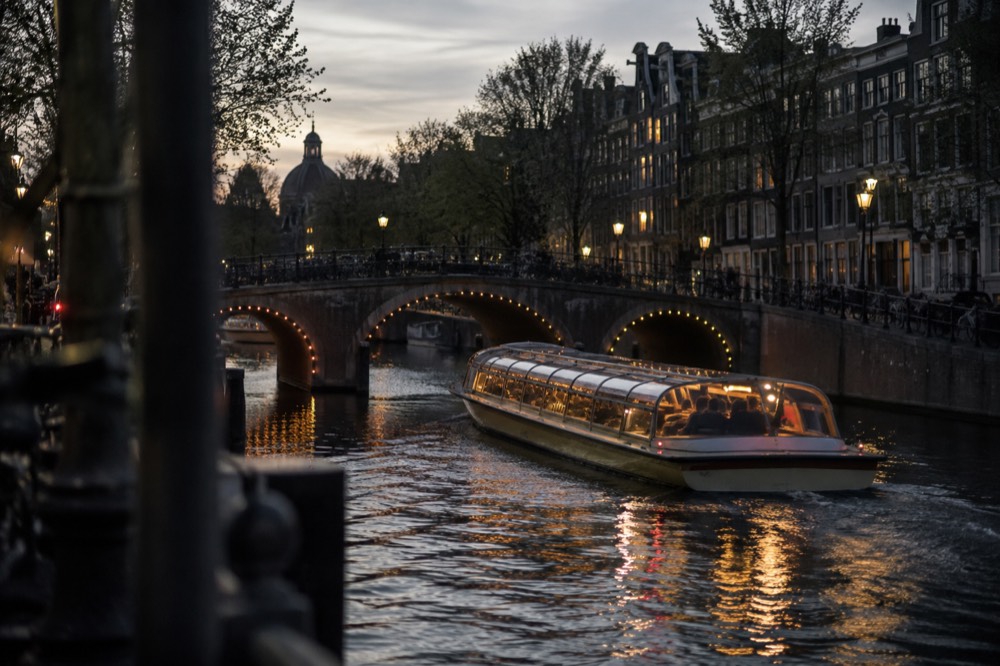 An illuminated canal cruise boat passing under a lit bridge in the evening