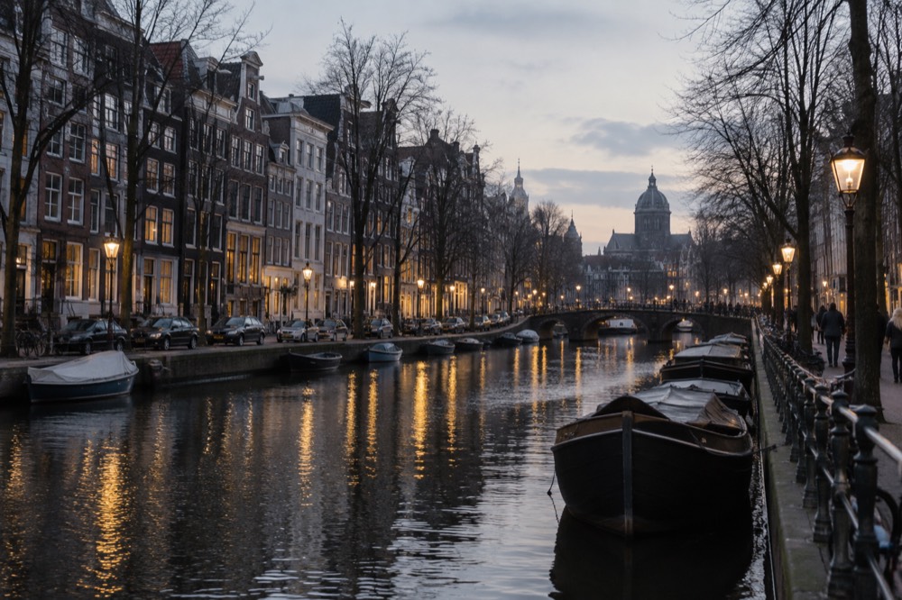 An evening view of a lit Amsterdam canal with boats moored along a dark water surface