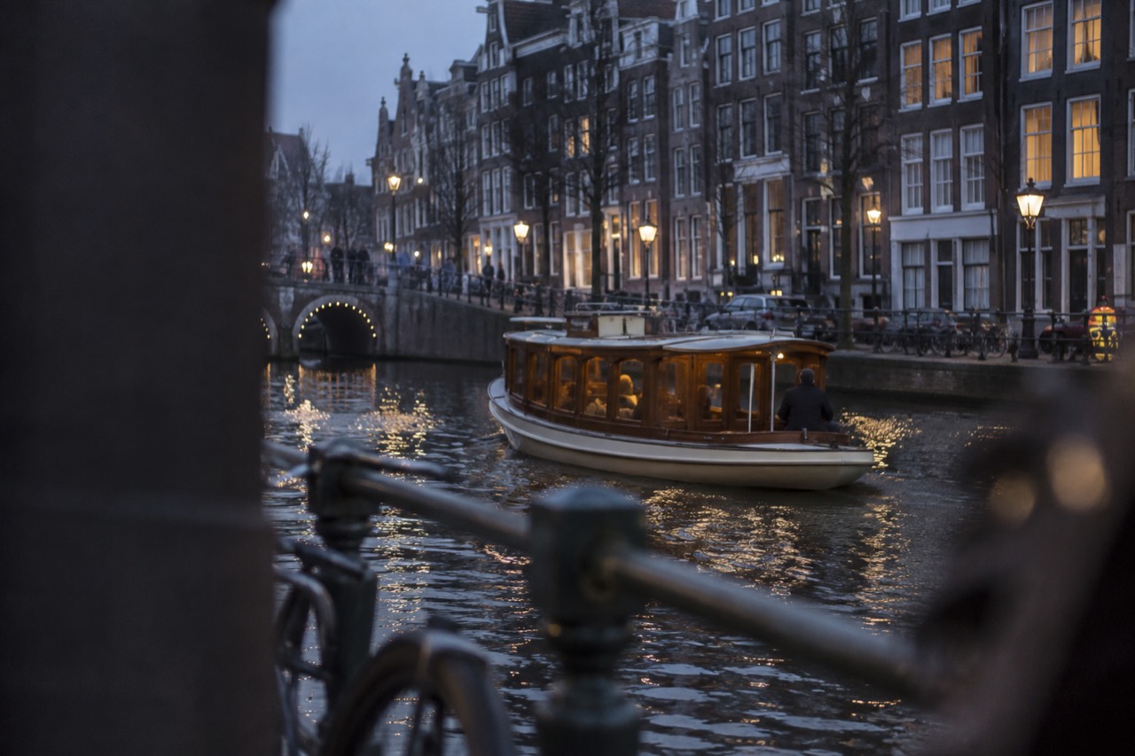 A classic wooden saloon boat cruising along an Amsterdam canal lined with narrow gabled houses