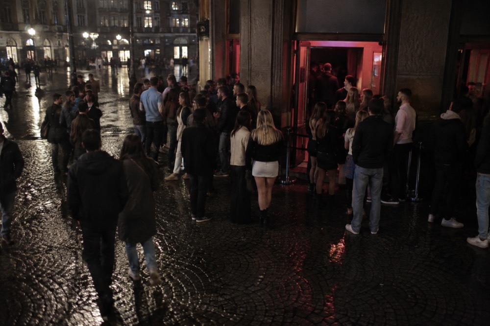 Crowd gathered outside a nightclub entrance in Amsterdam at night with neon signage glowing across the facade
