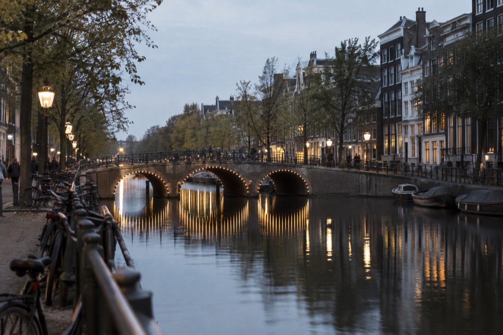 An evening canal in Amsterdam with illuminated bridge arches reflected in still water.