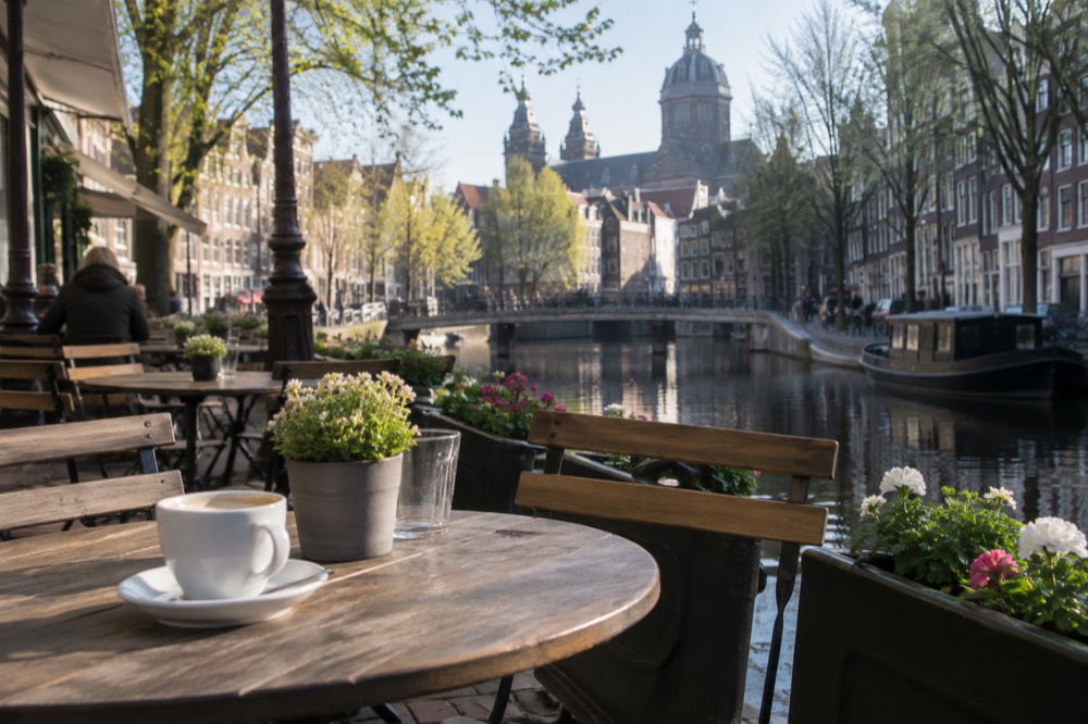 A canal-side terrace in Amsterdam with small tables, chairs and planters of flowers.