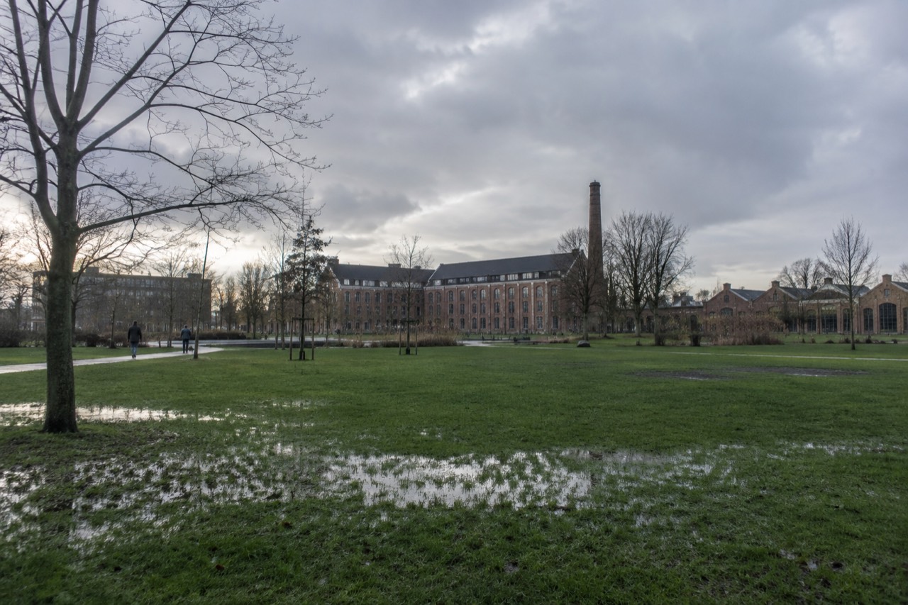 A wide green park lawn stretching toward a row of tall brick industrial buildings under a cloudy sky