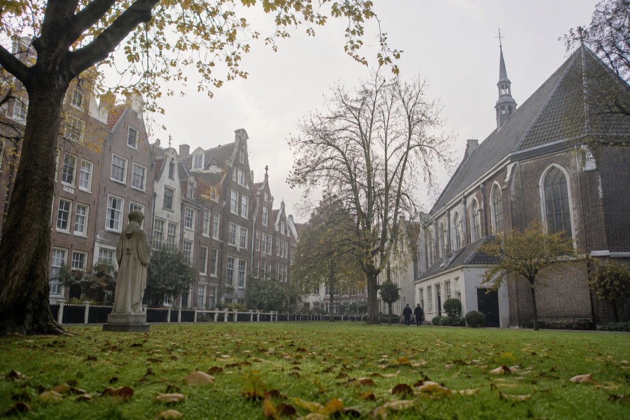A quiet enclosed courtyard in Amsterdam with tall narrow houses and a small chapel facing a green lawn