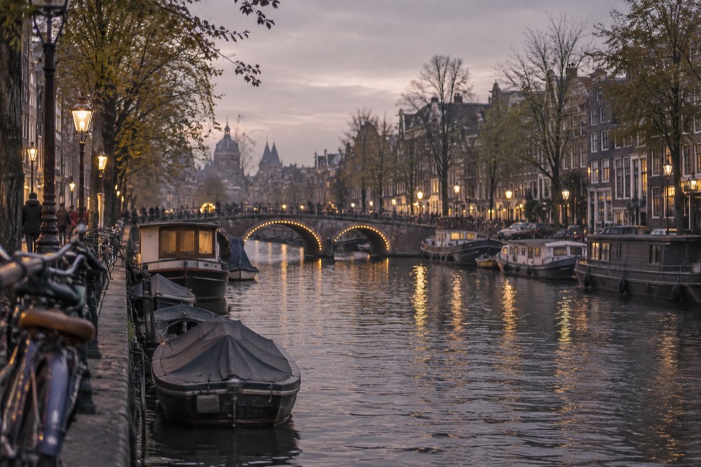 An Amsterdam canal at dusk with illuminated bridge and moored boats