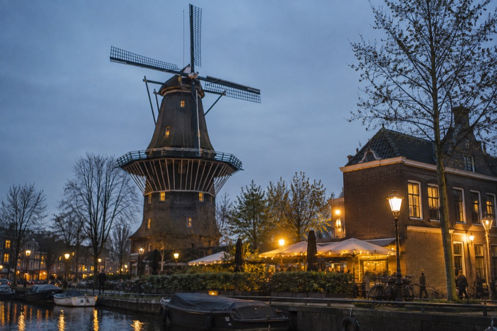 A tall wooden windmill with a gallery stage and four sails above brick buildings and bare trees