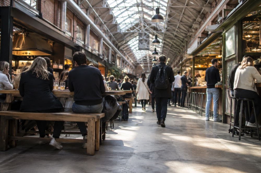 Interior of a busy food hall with food stalls and shared wooden tables under a glass roof