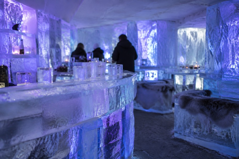 An ice bar interior with walls, benches and drinking glasses carved from ice, lit in blue and purple light