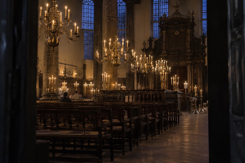 A vast candlelit synagogue interior with brass chandeliers hanging over rows of wooden benches