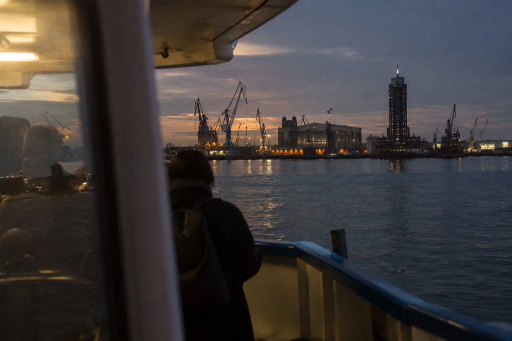 Ferry crossing open water towards a shipyard wharf with cranes and a tower on the far bank