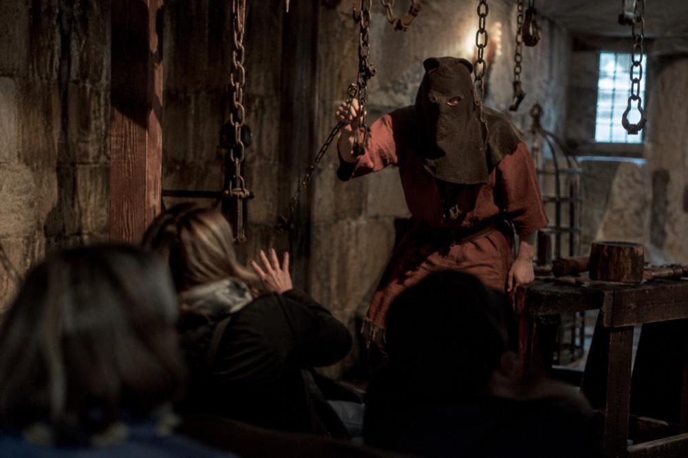 A dungeon actor mid-performance in a low-lit room with chains hanging from the ceiling and a startled visitor in the front row.