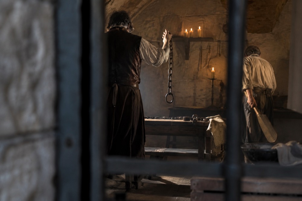 Two actors in period costume, one wielding a rusty chain, the other a wooden paddle, performing in a candle-lit torture room.