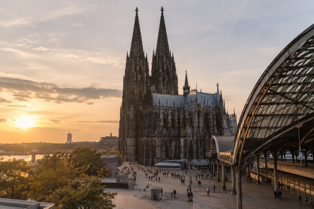 The gothic twin spires of Cologne Cathedral seen from a plaza with the main railway station roof at the edge of the frame