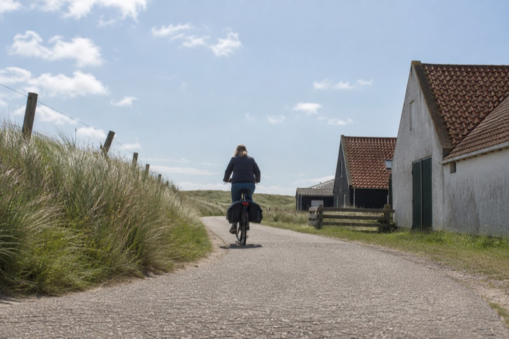 A cyclist on a flat path between dune grass and a row of low farm buildings