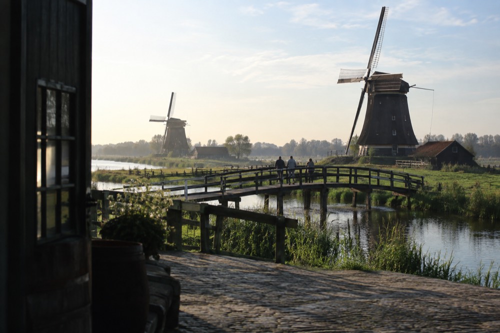Historic windmills beside a river with a wooden footbridge and green meadows
