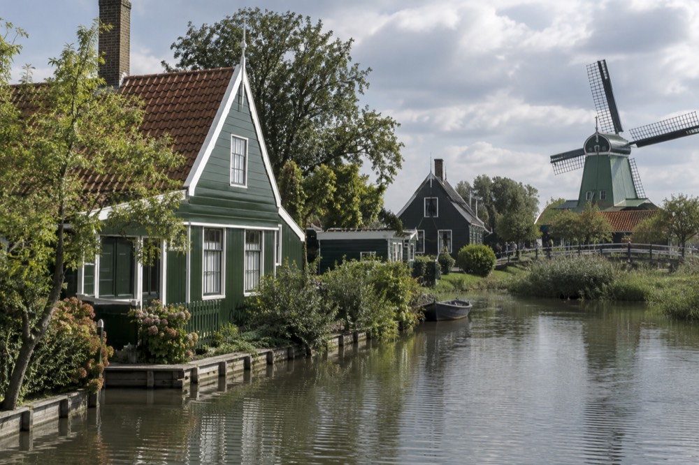 Green wooden historic houses along a quiet waterway at Zaanse Schans with a turning windmill behind them