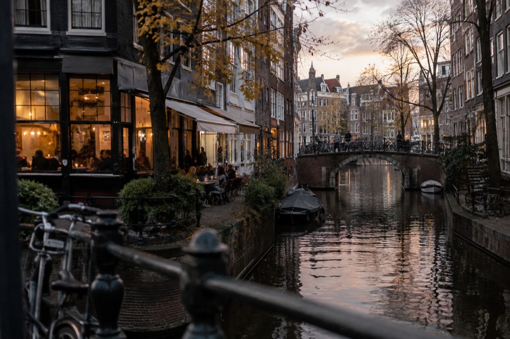 A narrow Amsterdam canal street at dusk with warm light glowing from café windows