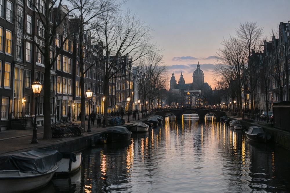 An evening view of a narrow Amsterdam canal lined with lit windows and old brick facades.