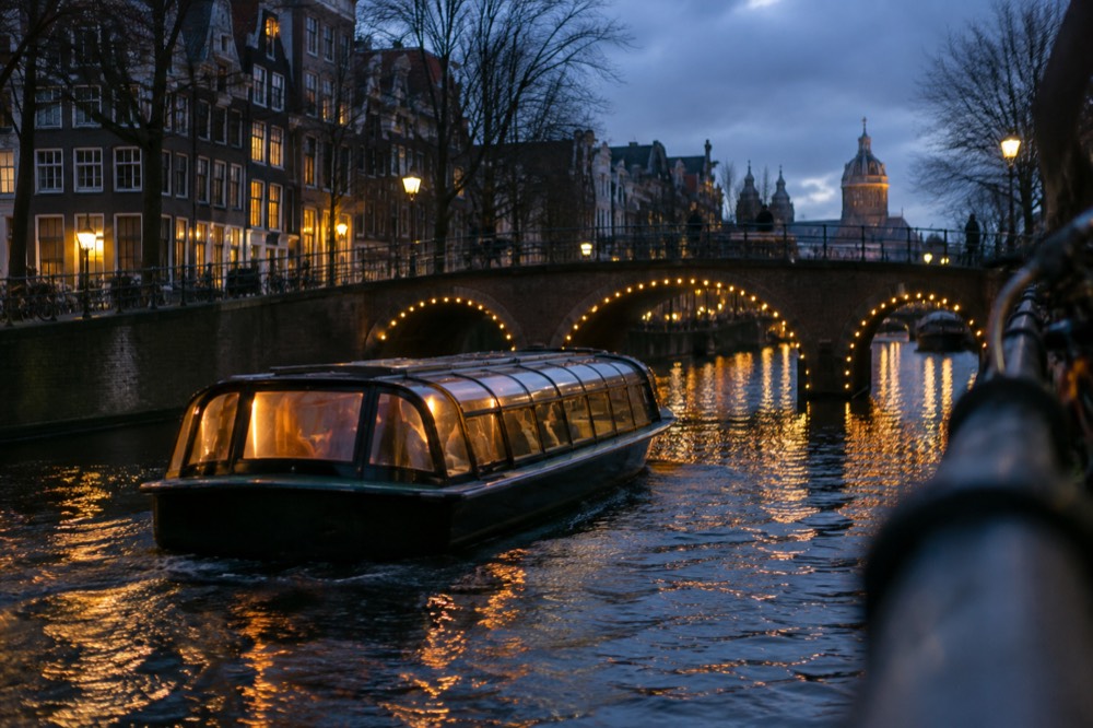 A covered canal boat passing under lit bridges on a dark evening