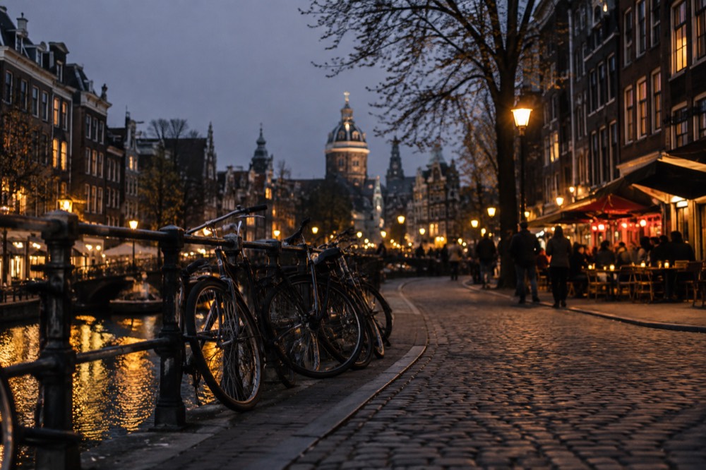 A historic Amsterdam street at dusk with cafe lights glowing and bicycles parked along the canal railing