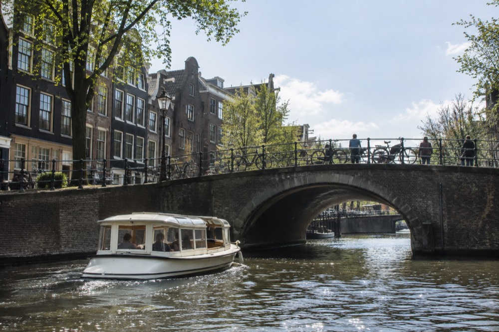 A small electric canal boat passing under a stone bridge with gabled houses behind