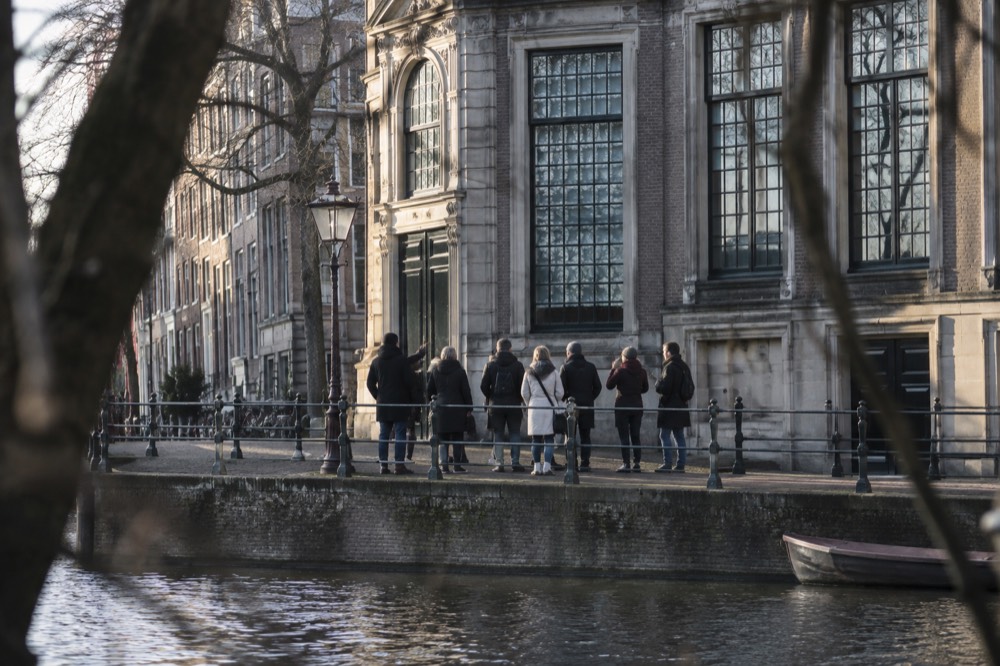 Walkers pausing at a canal-side building while a guide points out architectural details